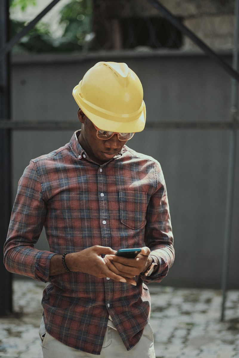 man in red blue and white plaid dress shirt wearing yellow hat with contractors insurance holding black smartphone